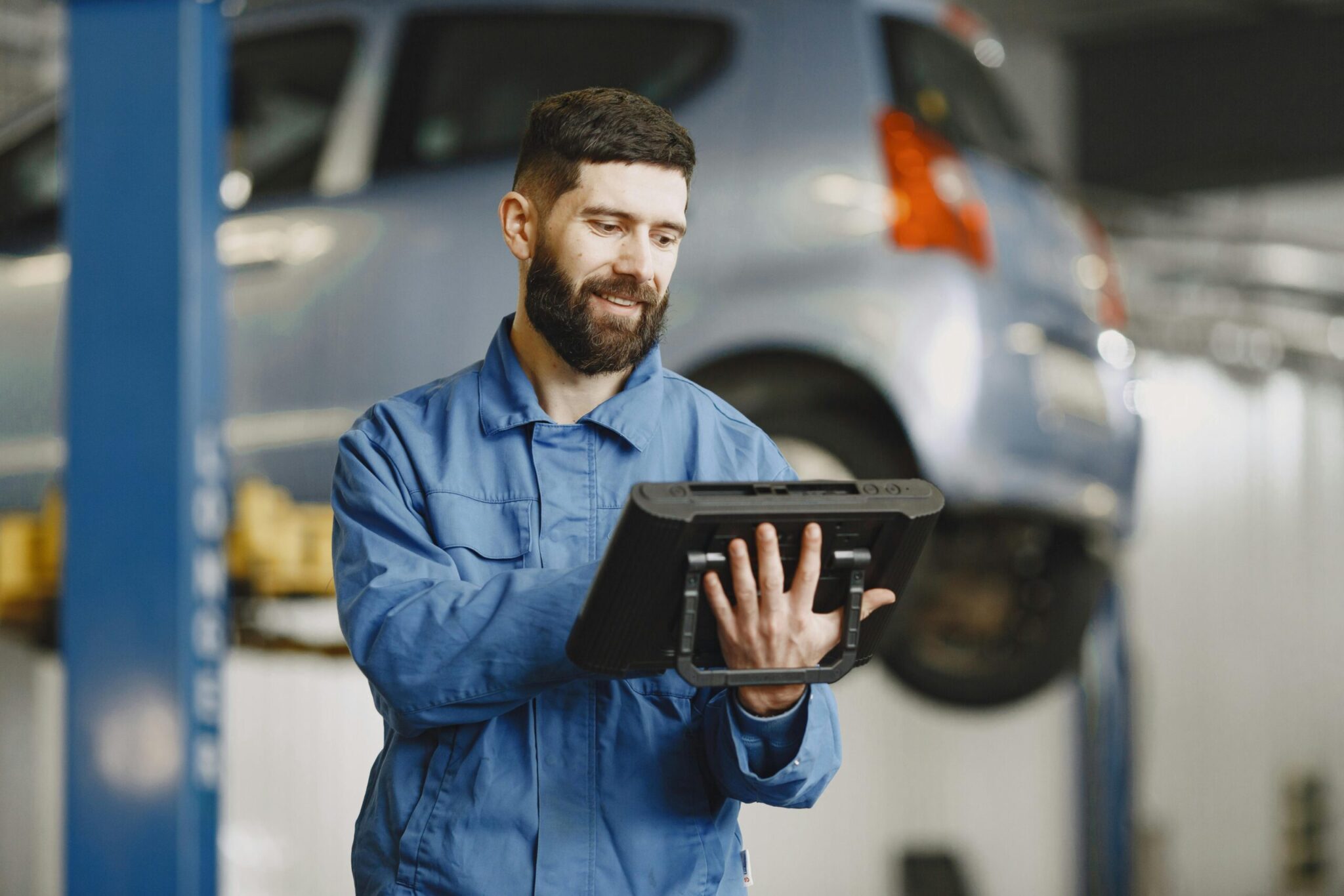 A mechanic in a blue jumpsuit diagnosing an electrical issue on a scan tool with a light blue hatchback on a lift in the background.